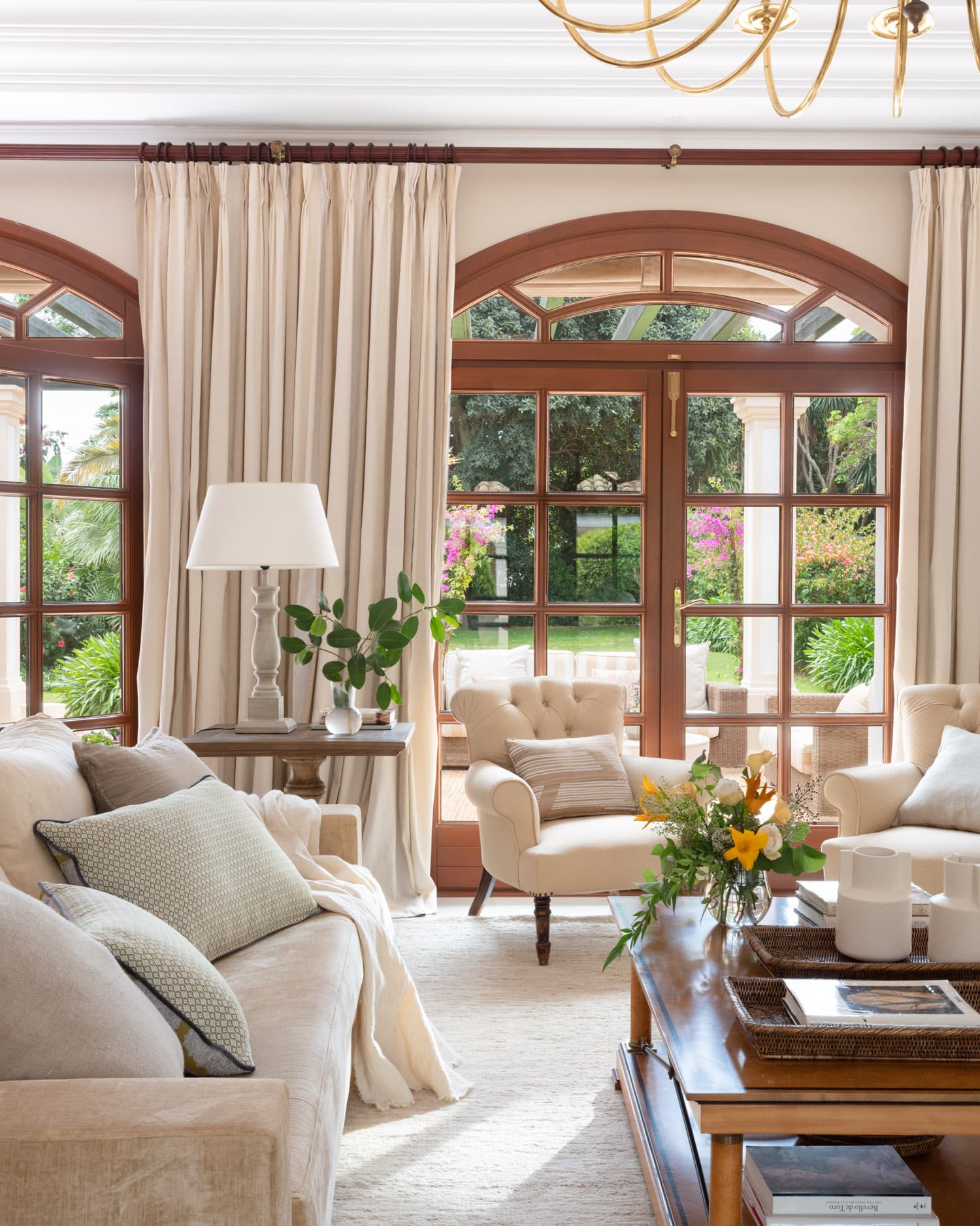Primer plano del salón principal mostrando los grandes ventanales de arco con carpintería de madera oscura y vistas a un jardín florido con buganvillas rosas. Un sofá de lino crema con cojines texturizados, un sillón capitoné y una mesa auxiliar de madera antigua conforman un rincón íntimo. Una luminaria esférica dorada aporta el toque de diseño contemporáneo.