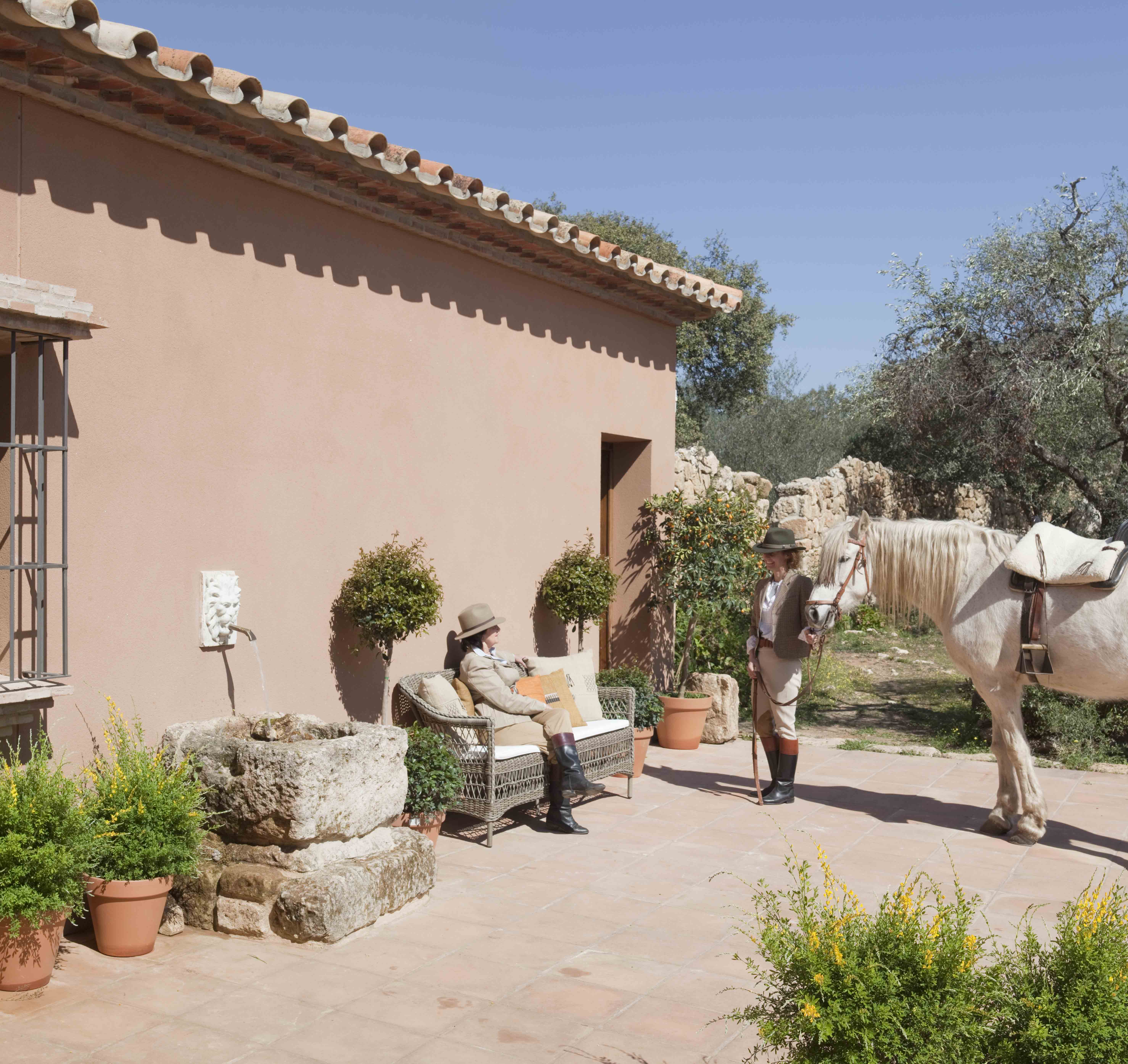 Imagen exterior de la finca con fachada en tonos tierra, donde dos personas con atuendo ecuestre —sombrero, botas y pantalones de montar— interactúan junto a un caballo blanco. La escena está enmarcada por árboles y macetas de barro, con el pozo de piedra antigua al fondo.