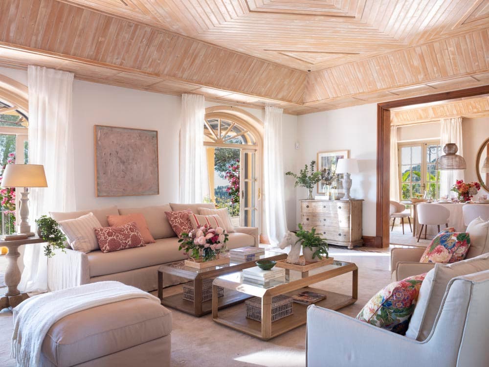 Amplio salón con un impresionante techo artesanal de madera clara en patrón geométrico, sofás en crema con cojines en rojo coral y floral, mesa de centro de madera y cristal, y una cómoda decapada. Al fondo se vislumbra el comedor a través de un arco.