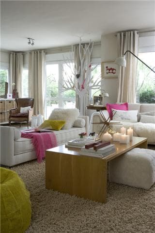 Vista alternativa del salón con sofás blancos en formación de rincón, el árbol de ramas blancas al fondo junto a las ventanas, mesita auxiliar en forma de seta y el puf verde lima en primer plano. El ambiente combina piezas de diseño con un carácter acogedor y espontáneo.