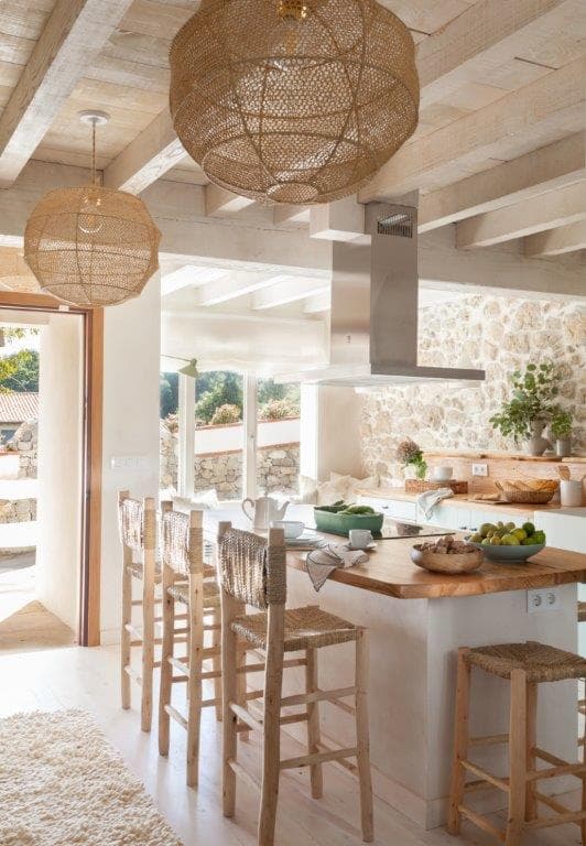 Cocina luminosa de planta abierta con gran puerta de madera al jardín, dos lámparas esféricas de mimbre colgantes y barra de desayuno con sillas de esparto. La combinación de materiales naturales y la abundante luz crean un ambiente cálido y acogedor.