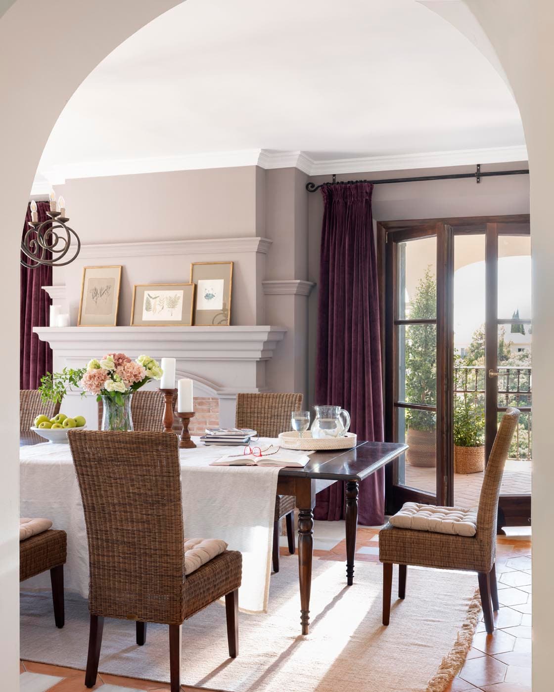 Comedor visto a través de un arco blanco, con una mesa oscura y sillas de mimbre rodeada de cortinas de terciopelo granate, una chimenea de mármol blanco y láminas botánicas enmarcadas. La luz natural que entra por las puertas francesas con vistas al jardín baña el espacio en calidez elegante.