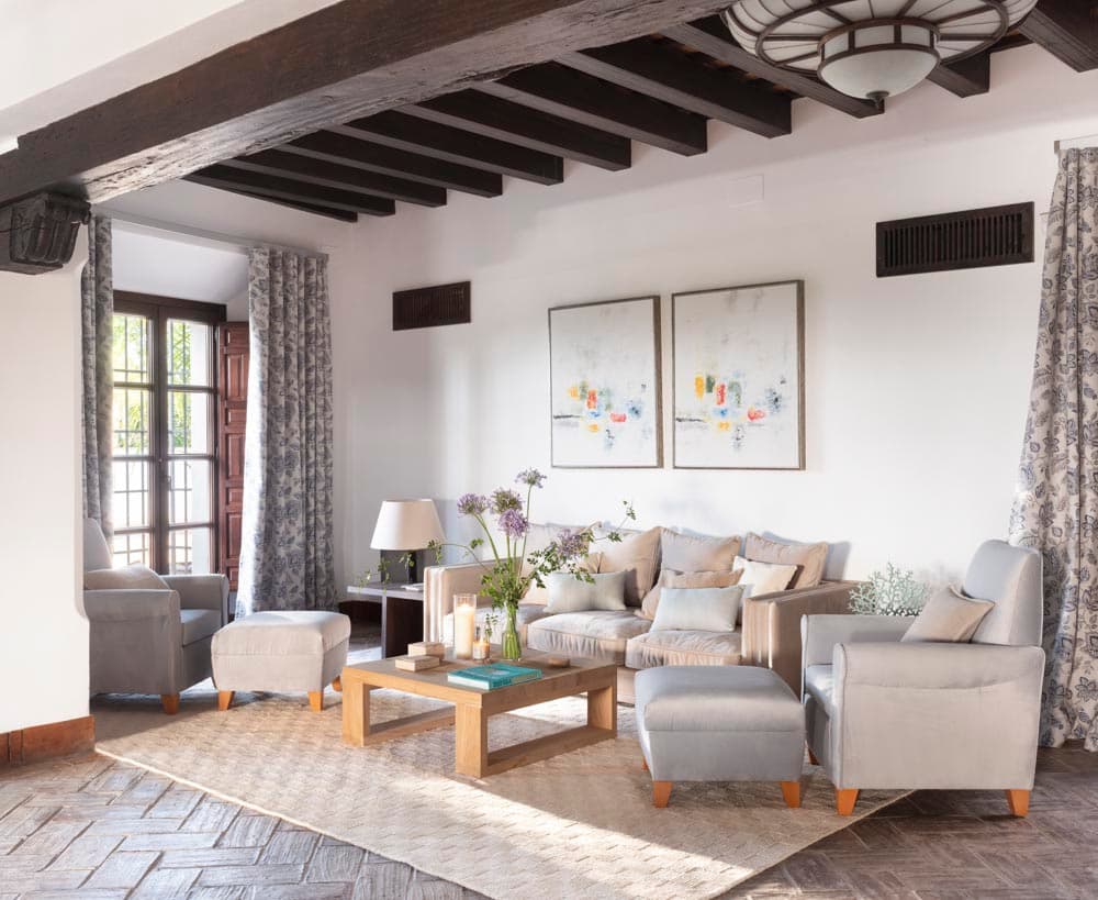 Sala de estar con sofá y butacas grises, mesa de centro de madera, pareja de cuadros abstractos coloridos sobre la pared y jarrón con flores de campo. Vigas de madera oscura al techo y cortinas estampadas con ventanas de reja.