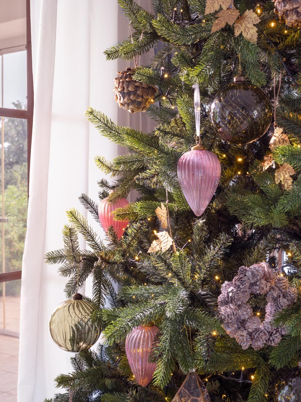 Acercamiento a las ramas del árbol de Navidad con adornos en vidrio soplado en tonos rosas y grises ahumados, piñas naturales y hojas doradas. La combinación de materiales naturales y ornamentos artesanales aporta profundidad y distinción a la decoración navideña.