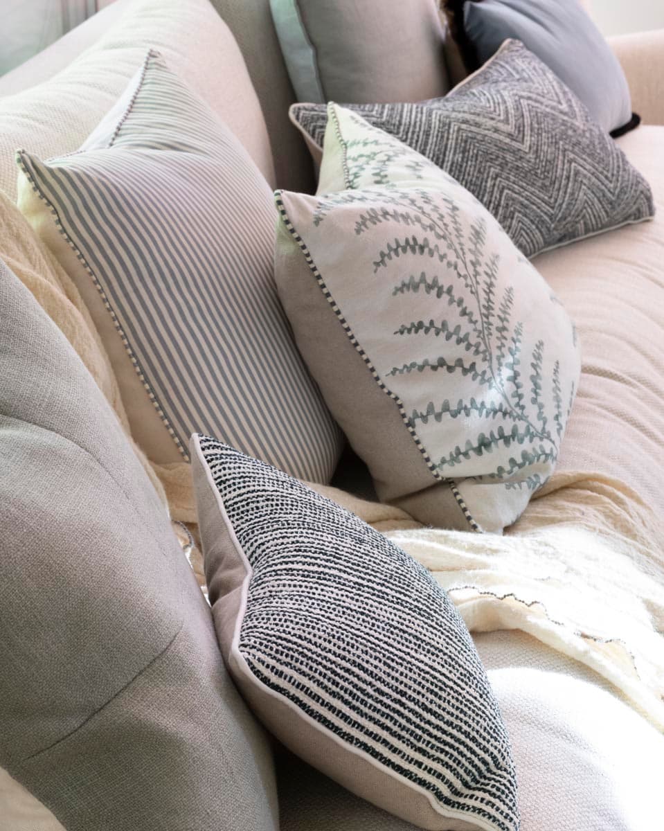 Primer plano de una composición de cojines sobre sofá lino en tonos crudo y gris: cojín rayado fino, cojín bordado con motivo de helecho, cojín en tejido de espiga oscuro y cojín chevron gris marengo, acompañados de una manta ligera en blanco roto. La variedad de texturas refleja el cuidado estilismo textil del proyecto.