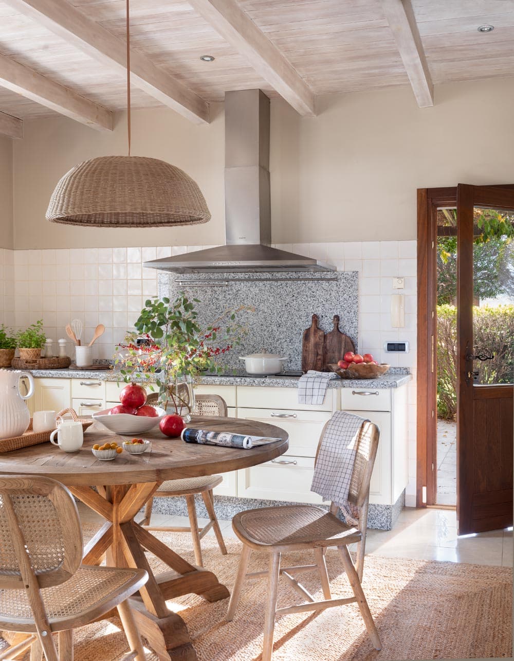 Cocina con puerta estable de madera con vidrio abierta al jardín exterior y luz natural entrante, mostrando la encimera de granito con accesorios decorativos, la lámpara de mimbre y parte de la mesa de desayuno con sillas de rejilla. La integración interior-exterior define el carácter mediterráneo de la vivienda.