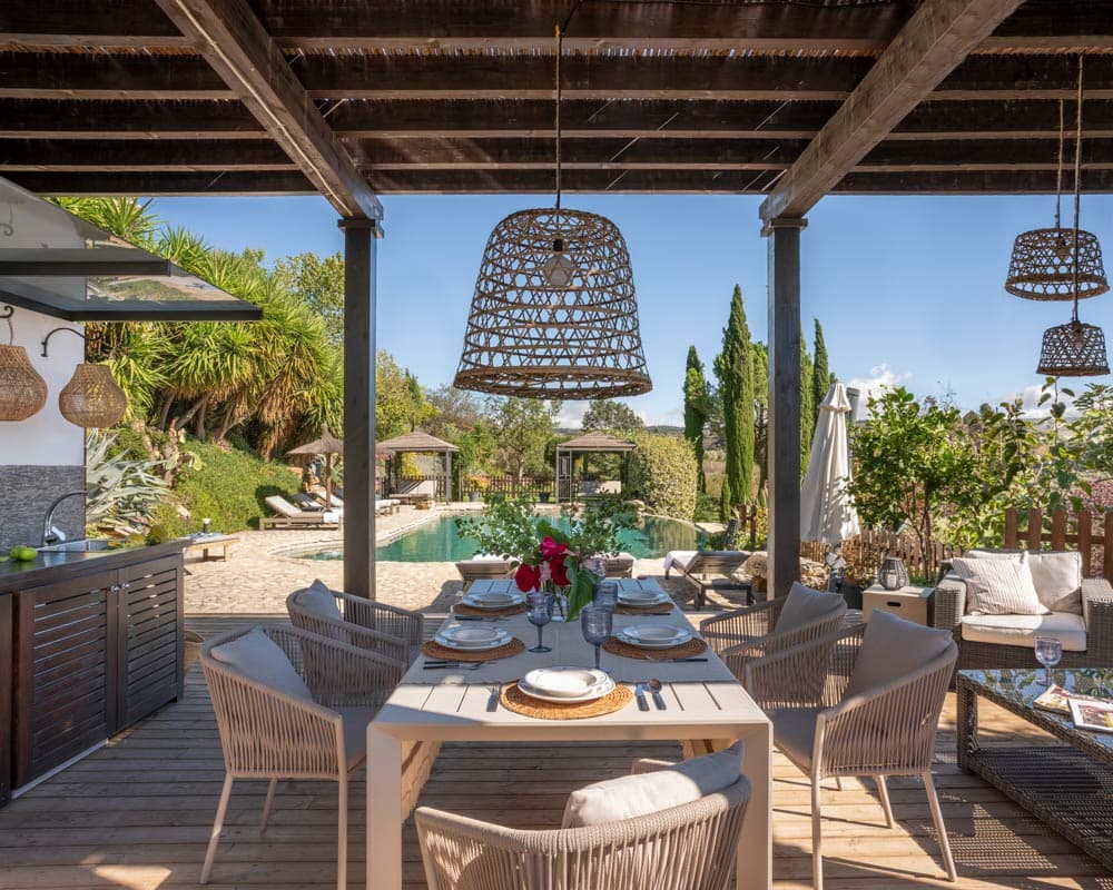 Mesa preparada bajo la pérgola con lámparas de ratán colgantes, mantel de lino, vajilla y flores rojas. Las sillas de trenzado y la cocina exterior integrada se muestran junto a una piscina y jardín al fondo.