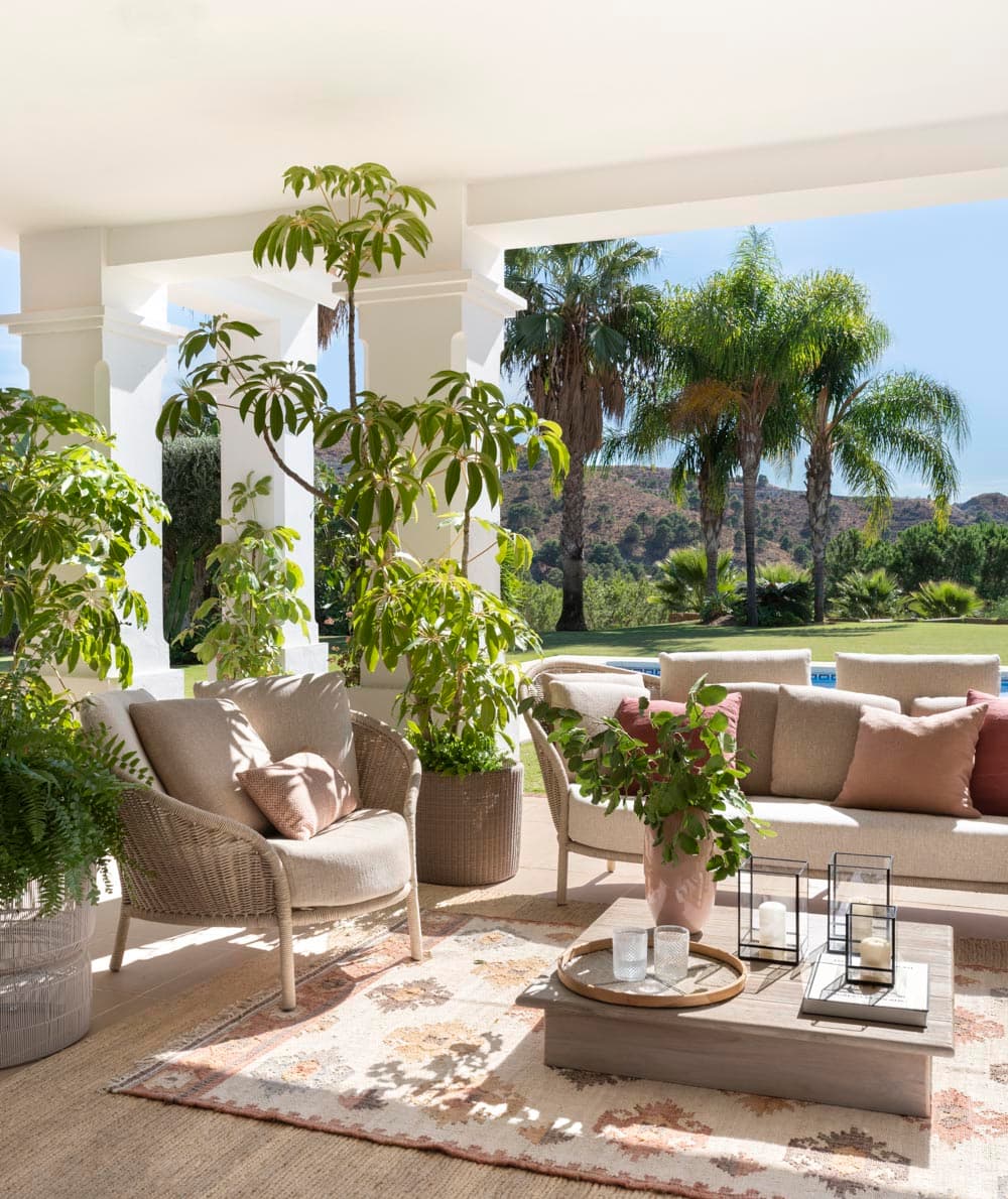 Zona de estar exterior con sillones de ratán en beige y sofá exterior, rodeados de vegetación exuberante: schefflera, ficus y plantas de jardín. Una jarrón de cerámica rosa con flores frescas centra la composición sobre la mesa de piedra. Al fondo se vislumbra la piscina, el jardín y palmeras bajo el cielo mediterráneo.