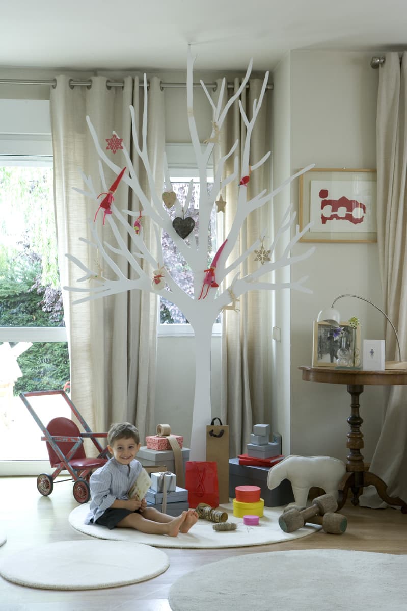 Vista amplia del salón con el árbol blanco decorativo al fondo, un niño sonriente sentado en la alfombra rodeado de regalos coloridos. La escena transmite la calidez navideña del hogar, con cortinas de lino natural y mesa auxiliar torneada.
