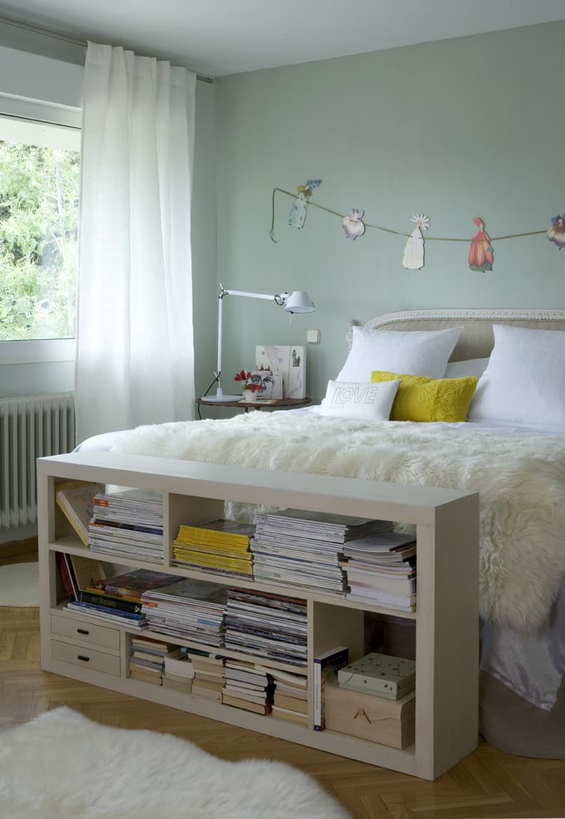 Vista lateral del dormitorio con paredes en verde menta, cama con ropa de cama blanca y cojín amarillo 'LOVE', estantería baja blanca repleta de revistas y libros. La pared del cabecero luce una guirnalda de ilustraciones de flores recortadas. Suelo de parqué y alfombra de pelo blanco.