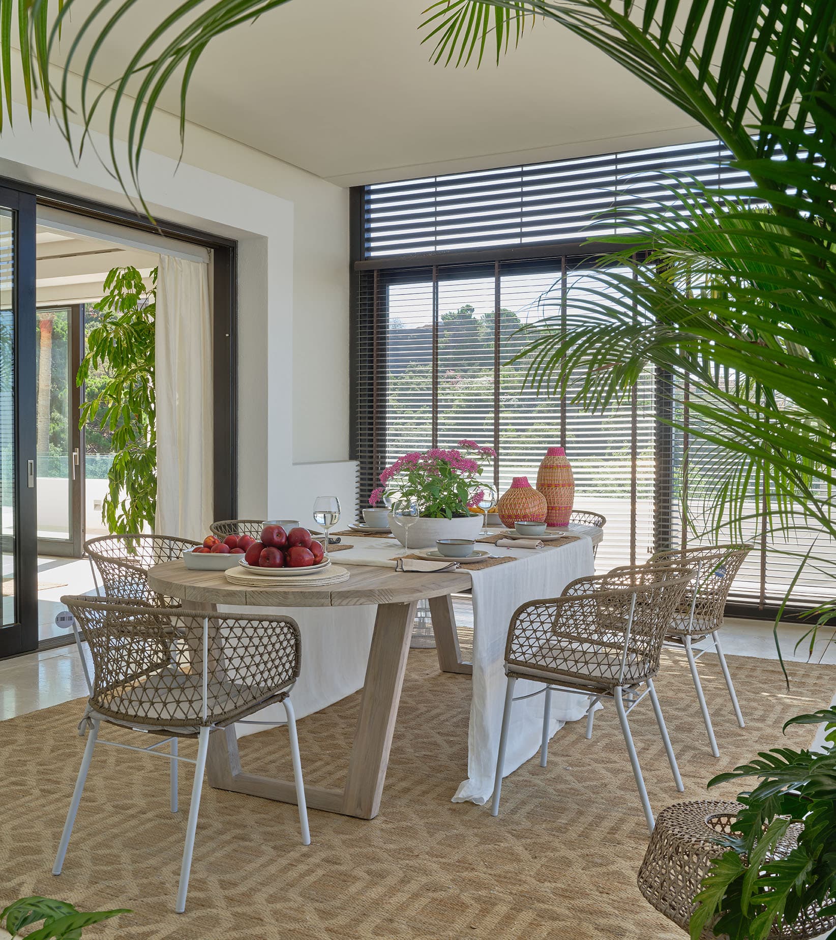 Comedor exterior cubierto con mesa de piedra reconstituida, sillas de metal y cuerda, decoración en terracota y rosa, rodeado de vegetación y persianas de lamas.