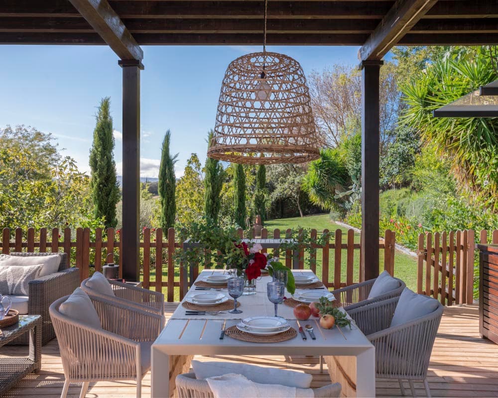 Mesa de comedor exterior servida bajo pérgola de madera con una lámpara de ratán grande colgante y vistas al jardín verde con cipreses y palmas. Las sillas de cuerda y la vajilla sobre mantel blanco aportan elegancia casual.