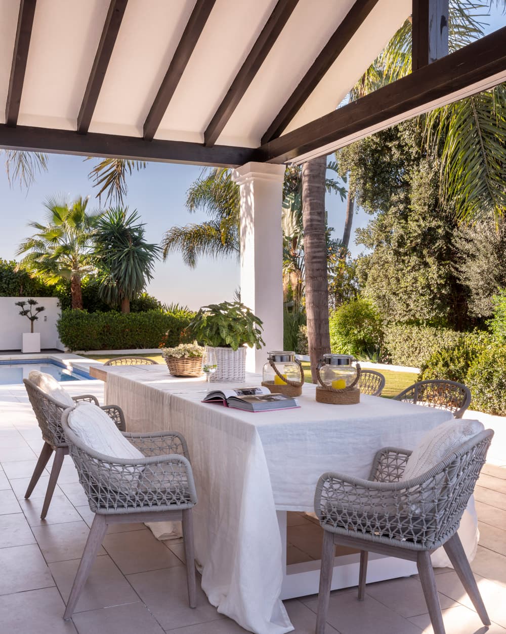 Mesa de comedor exterior con mantel blanco de lino, sillas de trenzado gris y jarras de limonada, bajo una pérgola con vigas oscuras con vistas a la piscina y a la vegetación tropical del jardín. El ambiente tropical y luminoso evoca el lujo mediterráneo al aire libre del cortijo.