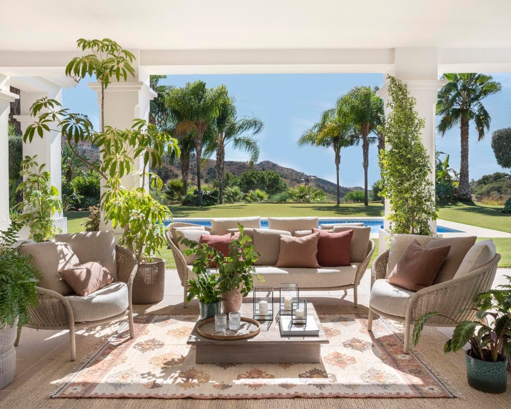 Amplio salón exterior bajo porche con mobiliario de ratán en beige y sofá curvo con cojines en terracota y rosa viejo. Mesa de centro rectangular de piedra con bandeja y velas. Las columnas blancas y la vegetación exuberante —palmeras, ficus, enredaderas— enmarcan la piscina al fondo con vistas a la montaña.