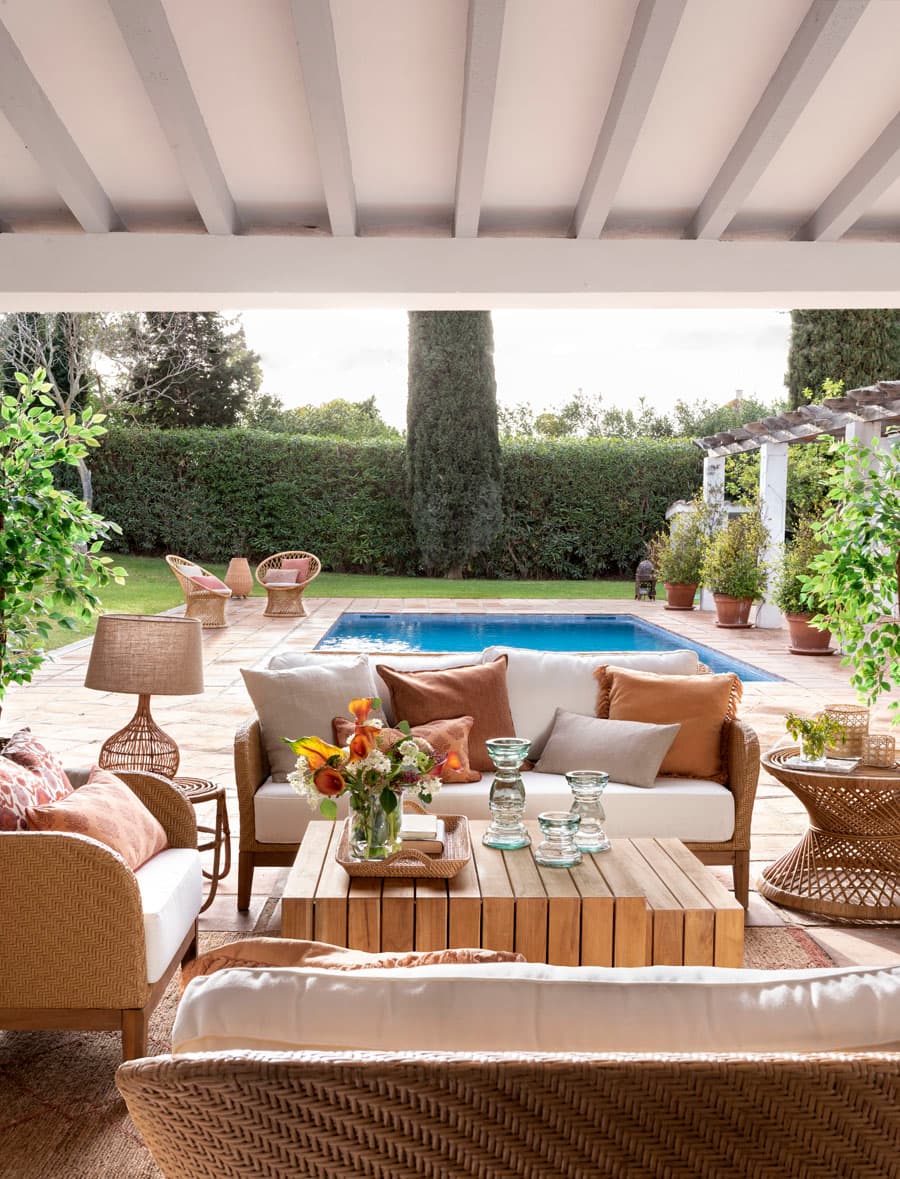 Vista destacada del porche con la zona de estar en primer plano y la piscina rectangular al fondo, enmarcada por setos de boj y vegetación mediterránea. Los sofás y butacas de ratán con cojines en tonos terracota, cobre y beige rodean una mesa de centro de madera con una bandeja de ratán que sostiene flores de temporada y portavelas de vidrio. Una lámpara de pie de ratán ilumina el rincón con calidez. A la derecha, una mesa auxiliar de ratán redonda completa la composición.