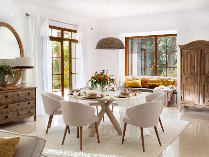 Comedor luminoso con mesa redonda de base cruzada en madera natural, sillas tapizadas en beige claro, lámpara de cestería colgante y un armario antiguo de madera noble junto a la cómoda. Los cojines amarillos mostaza y la ventana-banco con cojines complementan la calidez del ambiente.