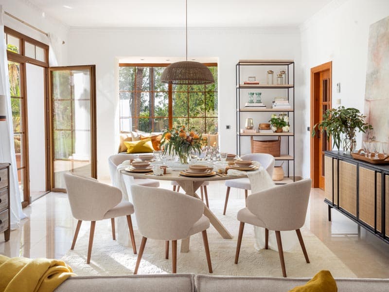 Vista amplia del salón-comedor con mesa redonda vestida, puertas francesas a doble altura y aparador negro con frontal de ratán, bañado por la luz natural del jardín mediterráneo.