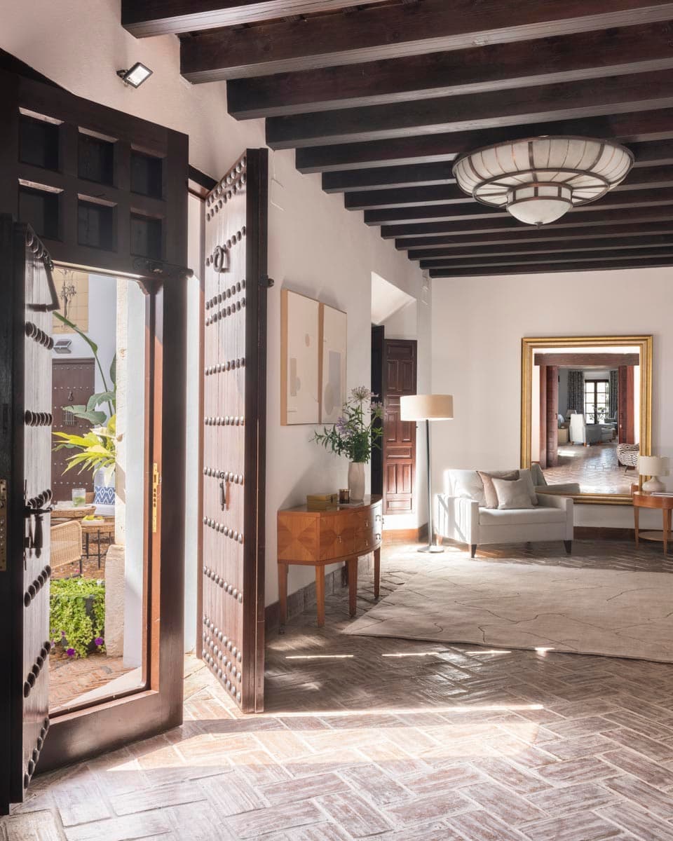 Zaguán de entrada con gran puerta de madera tachonada entreabierta que deja entrar la luz del patio. Aparador de madera, lámpara de techo articulada, sillón blanco al fondo y espejo dorado. Suelo de barro en espiga y vigas oscuras.