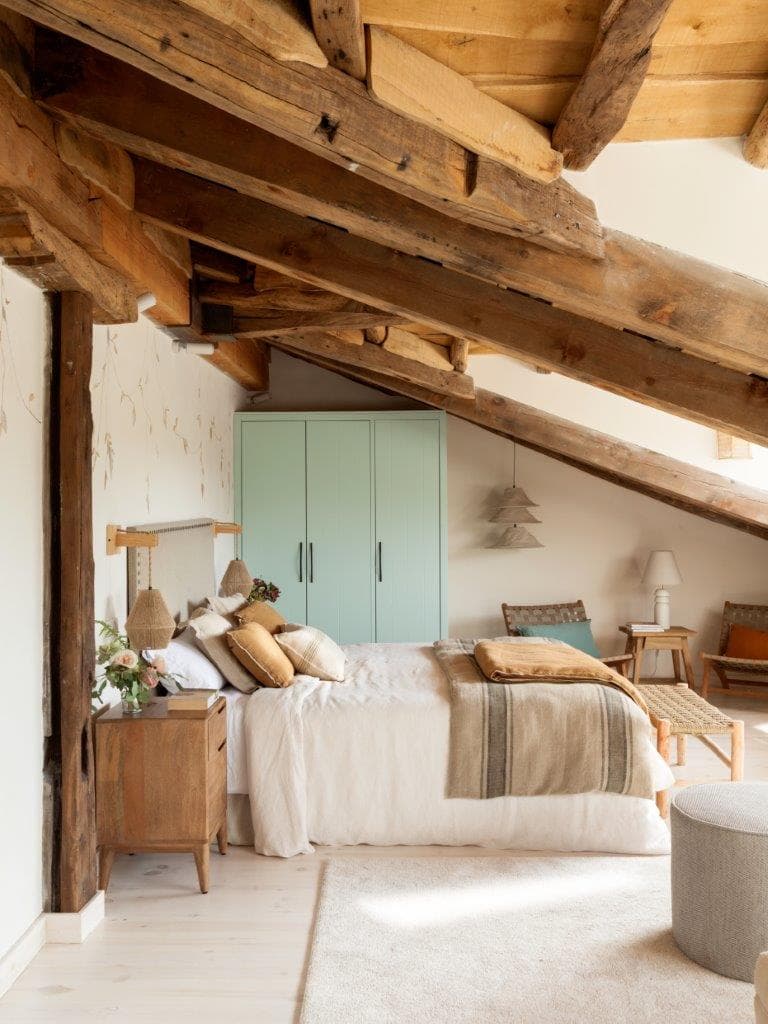 Vista alternativa del dormitorio en buhardilla con techos de vigas rústicas, cama con cabecero tapizado y tonos camel y lino, y armario verde menta empotrado. Rosas rosas y helechos en jarrón sobre la mesita añaden frescura al espacio.