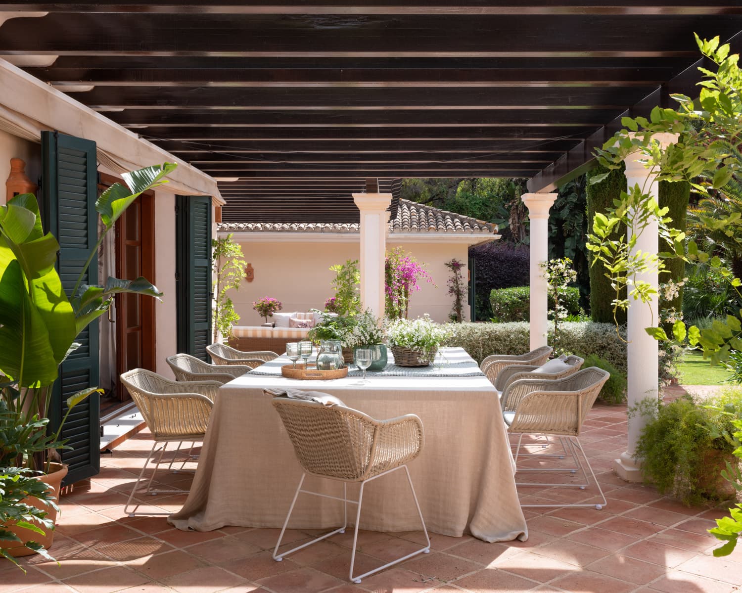 Generoso comedor al aire libre bajo una pérgola de madera oscura sostenida por esbeltas columnas blancas clásicas. La gran mesa rectangular, cubierta con mantel de lino natural, está rodeada de sillas de fibra trenzada. Puertas con contraventanas verdes y vegetación tropical enmarcan la estancia, creando un ambiente de elegancia mediterránea.
