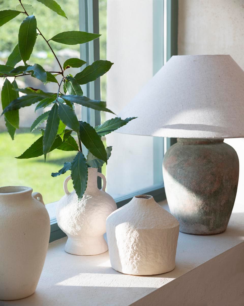 Composición sobre alféizar de ventana verde con jarrones artesanales de cerámica en blanco rugoso y gres envejecido verde, lámpara de sobremesa con base de cerámica antigua y rama de árbol fresca.