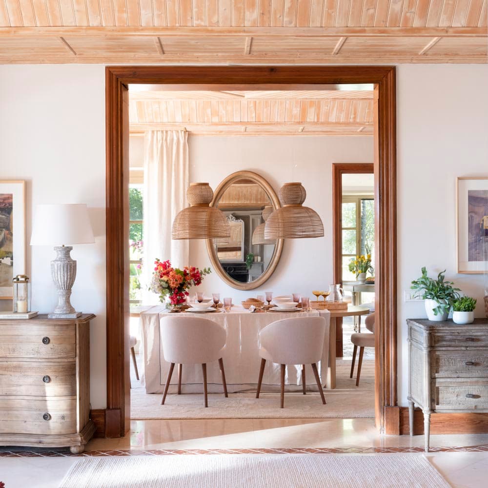 Encuadre desde el salón a través de un arco de madera oscura que enmarca el comedor, con cómodas envejecidas a ambos lados y una mesa oval bajo lámparas de ratán. El techo artesanal de madera clara y los cuadros abstractos en tonos rosados refuerzan la calidez del conjunto.