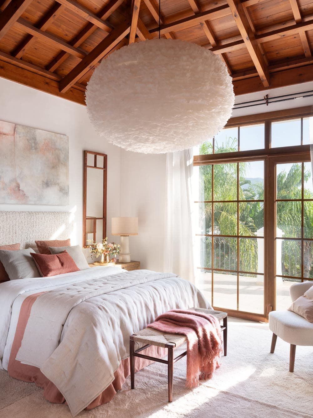 Dormitorio principal con impresionante lámpara colgante esférica de plumas blancas bajo un techo de vigas de madera, cama vestida en tonos blancos y rosa palo con cabecero textil, y gran ventanal de madera con vistas a palmeras tropicales. El conjunto combina lo orgánico y lo natural con una estética de lujo tranquilo.