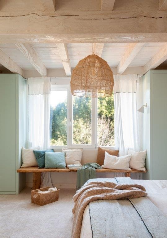 Dormitorio con banco corrido de madera bajo ventana con armarios integrados en verde menta a ambos lados, cojines en tonos azul y verde menta y lámpara colgante de rafia. La vista al jardín y la luz natural crean un espacio sereno y luminoso.