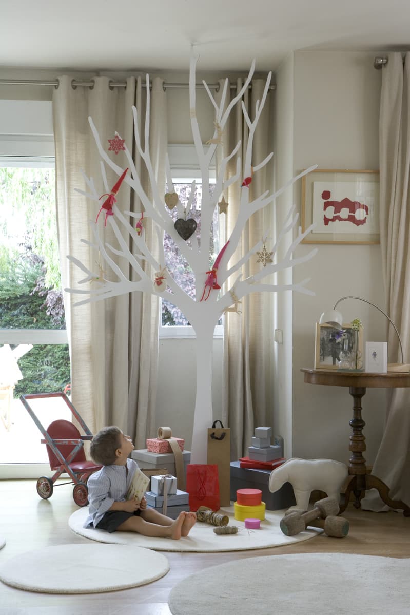 Niño sentado en el suelo frente al gran árbol blanco decorativo, rodeado de regalos de Navidad sobre una alfombra redonda. El espacio luminoso combina cortinas de lino, arte enmarcado y una mesa auxiliar torneada de madera oscura.
