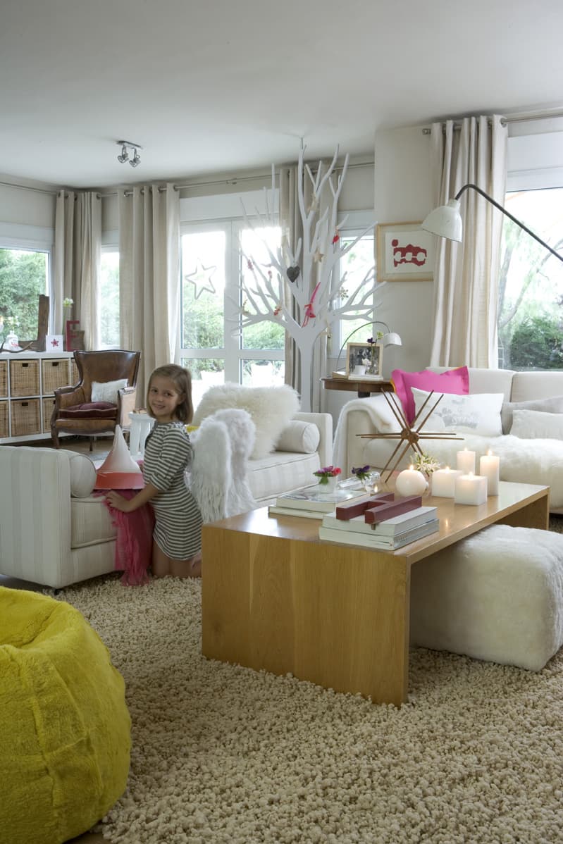 Escena del salón principal con la niña de pie junto al sofá blanco, sujetando un objeto decorativo. La composición muestra con claridad la mesa de centro de roble con la decoración navideña —velas encendidas, libros de arte y una bandeja roja—, el sillón blanco con cojín de piel y el gran árbol decorativo blanco al fondo. El sofá de gran formato blanco con cojín fucsia y fucsia sobre el brazo se aprecia a la derecha, junto a la lámpara de suelo negra y el puf amarillo en el ángulo inferior. Las ventanas inundan el espacio de luz exterior.