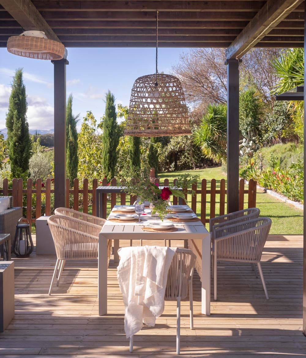 Mesa de comedor exterior preparada bajo pérgola de madera con lámpara de ratán colgante y vistas al jardín con cipreses. Las sillas de trenzado y el jardín bien cuidado con rosas crean un ambiente elegante para comer al aire libre.