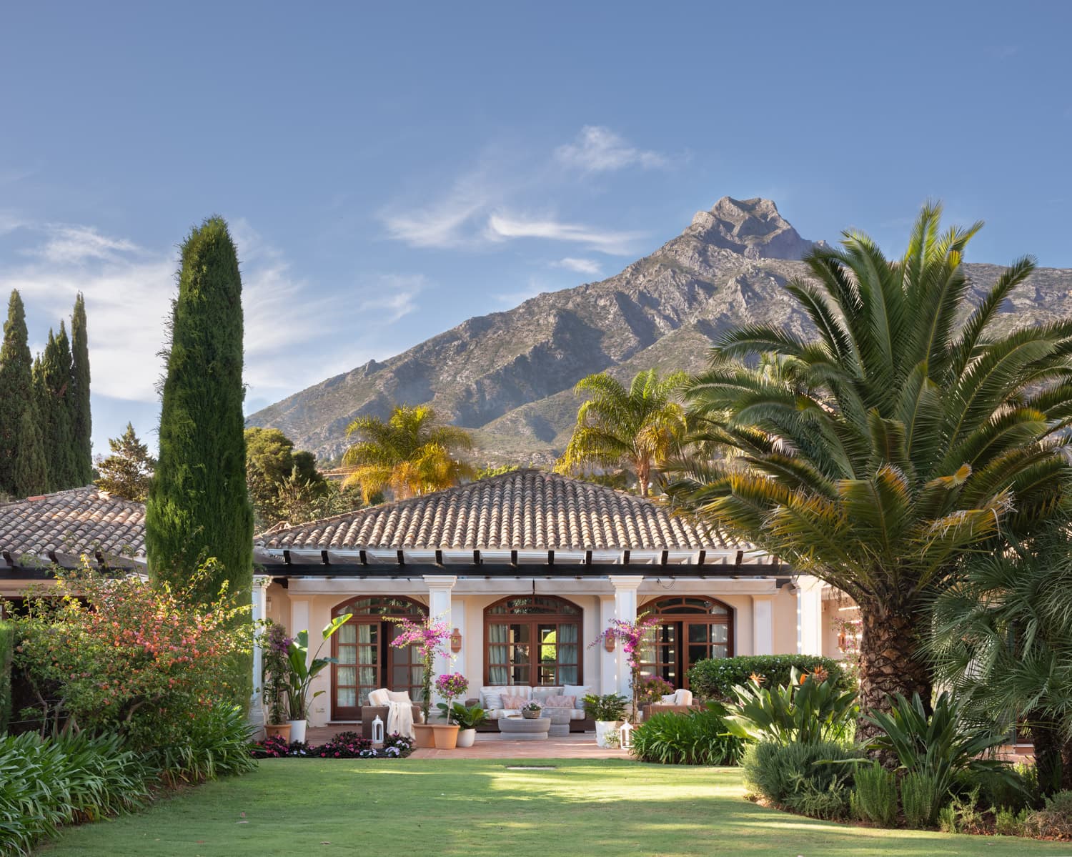 Vista exterior de la villa Capellanía desde el jardín. La fachada de estilo mediterráneo con tejado de teja árabe y porche con puertas francesas de madera aparece rodeada de una exuberante vegetación subtropical: palmeras, buganvillas en flor, cipreses y setos. Al fondo, la imponente silueta de La Concha o Jaén domina el horizonte bajo un cielo azul despejado.