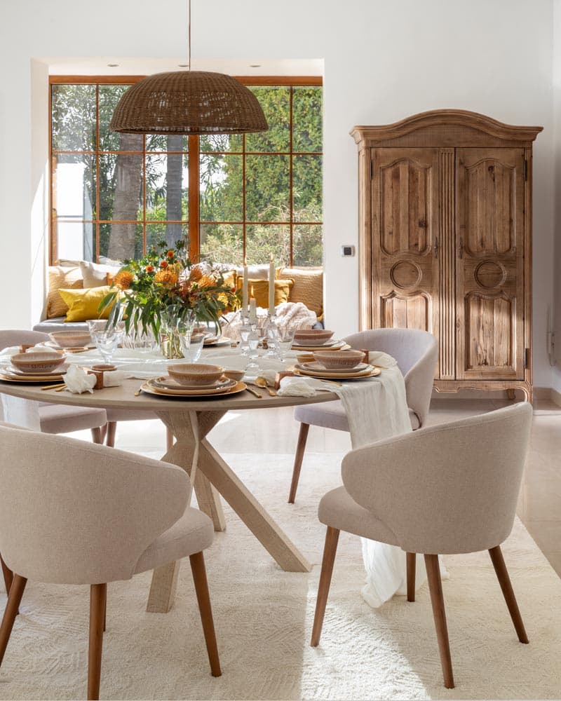 Detalle del comedor con mesa redonda de madera cruzada vestida para una ocasión, sillas en bouclé beige y un imponente armario colonial tallado en madera natural al fondo.