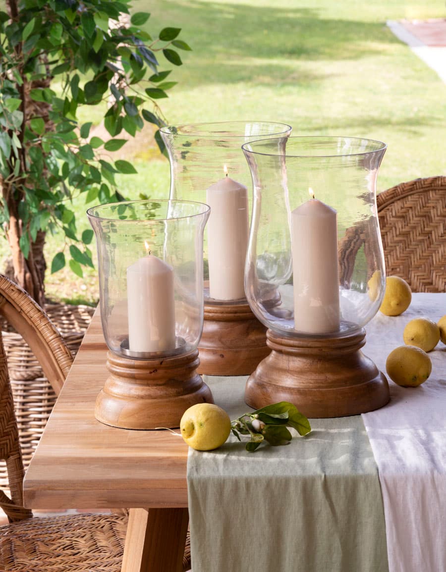 Vista de la mesa de comedor exterior con un elegante centro compuesto por tres portavelas de cristal soplado con base torneada de madera oscura, de diferentes alturas, con velas de pilar en tono crudo. El camino de mesa de lino verde salvia y los limones dispersos sobre la tabla de madera aportan frescura y naturalidad mediterránea. Al fondo, la apertura del porche al jardín con una silla de ratán y vegetación.