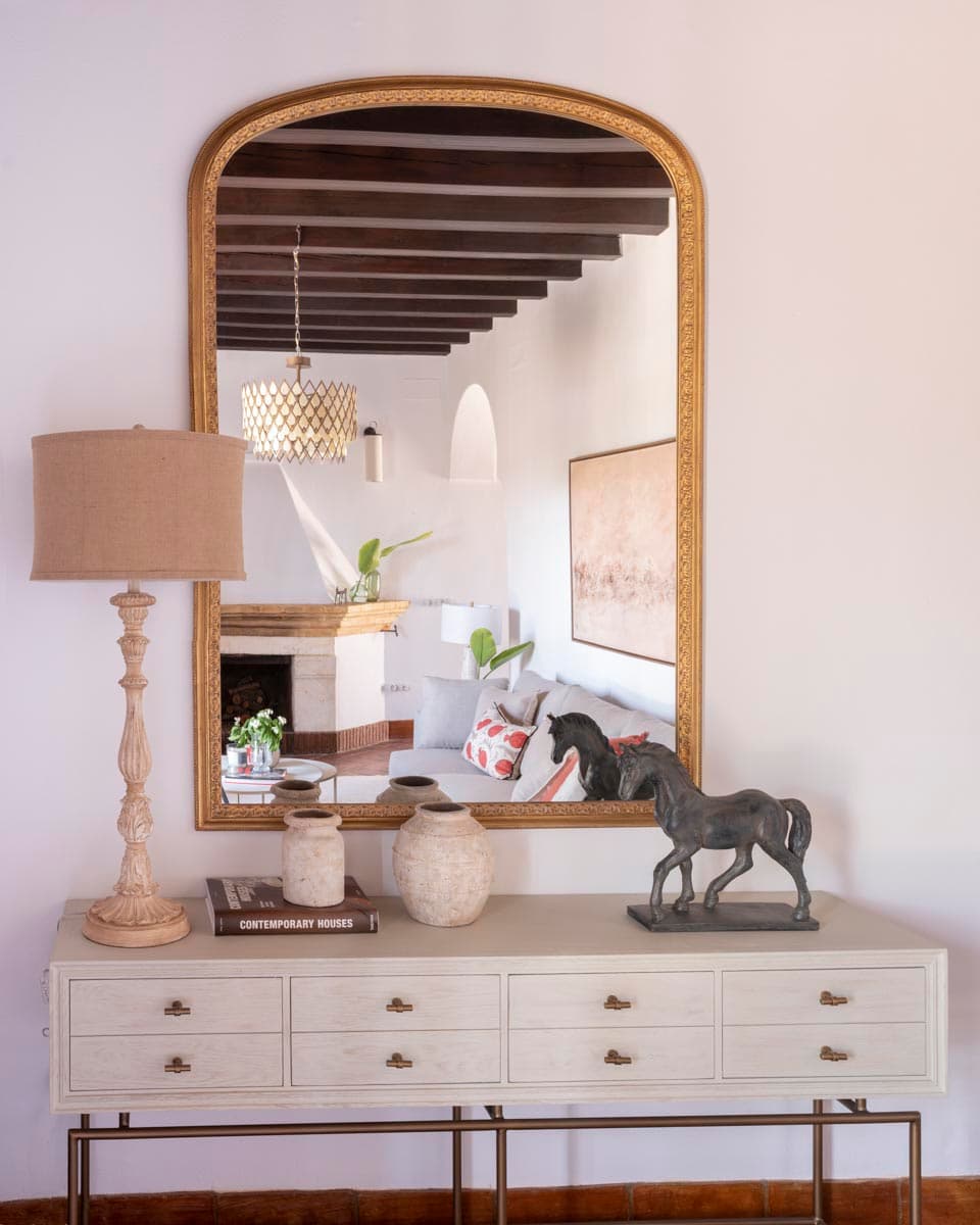 Detalle del aparador blanco de cajones con espejo de arco en marco dorado y escultura de caballo en bronce. Lámpara de sobremesa de madera tallada, jarrones de cerámica rústica y libro de arte sobre la superficie. Vigas y chimenea al fondo reflejadas en el espejo.