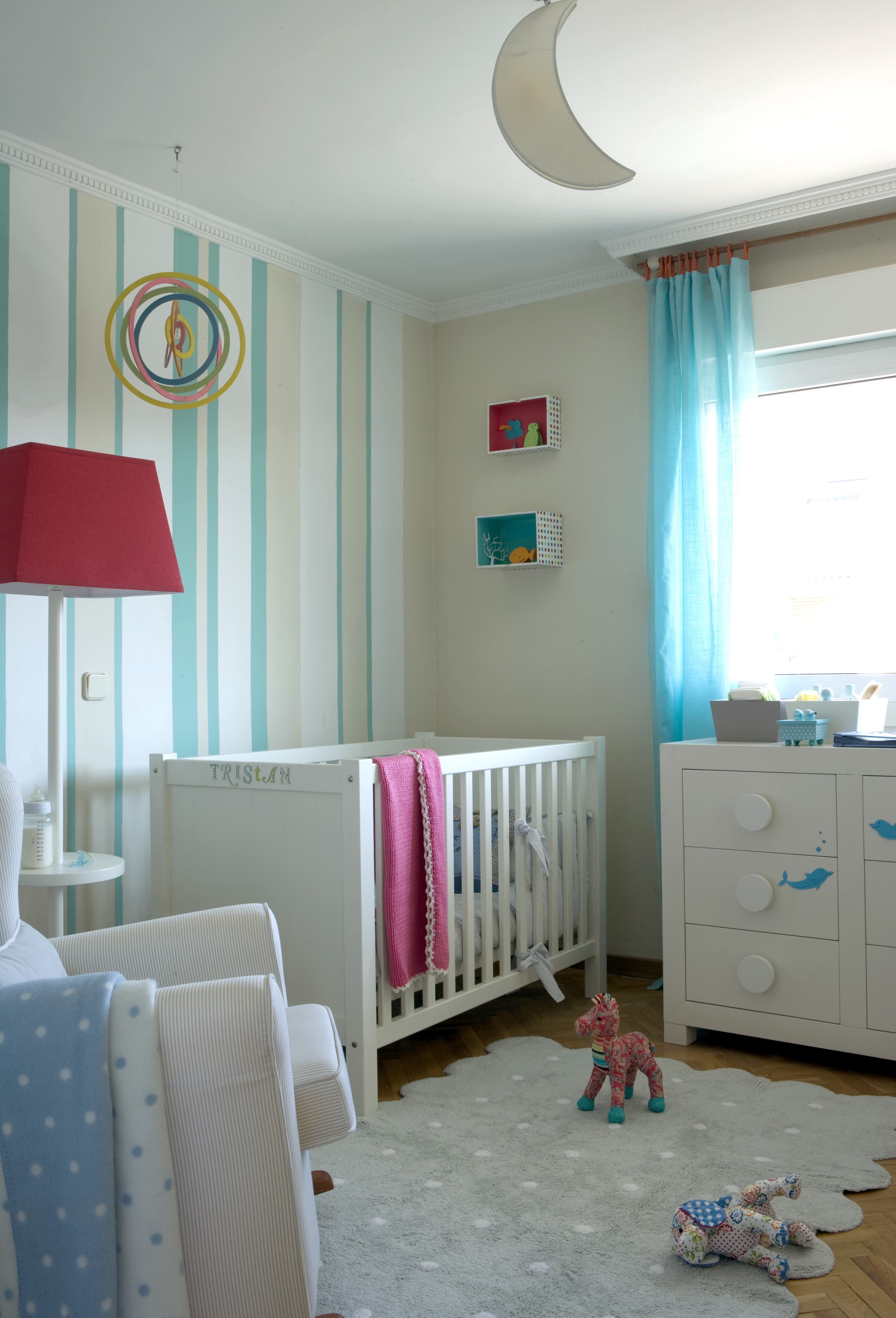 Habitación infantil de bebé con papel pintado de rayas verticales en turquesa y crema, cuna blanca con el nombre del bebé, cómoda con tiradores de círculo, lámpara de pie en rojo y lámpara de techo en forma de luna. Los elementos decorativos en turquesa y los peluches crean un ambiente tierno y estimulante.