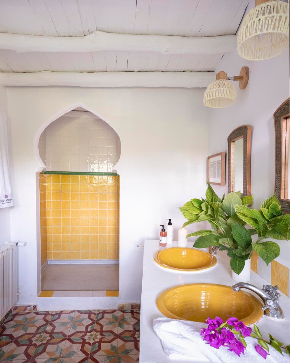 Baño de doble lavabo enmarcado por un arco de yeso blanco, con encimera blanca y lavabos artesanales amarillos, espejos de marco de mimbre y suelo de mosaico hidráulico. La ducha de azulejo azul marino se aprecia al fondo.