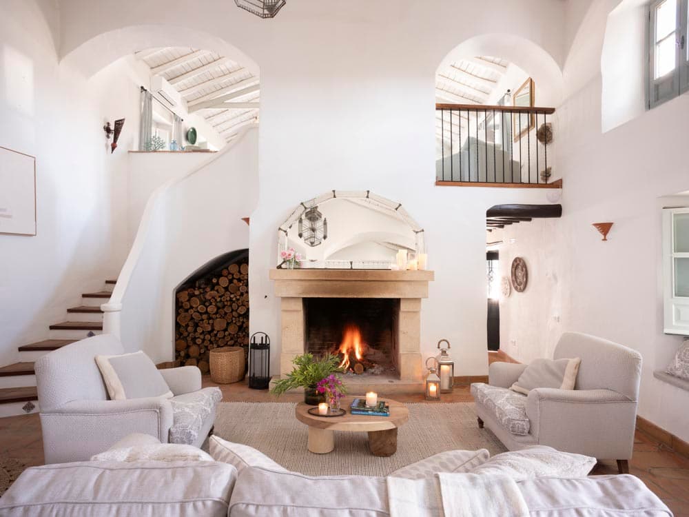 Salón principal de doble altura con chimenea de piedra encendida, leñera empotrada y espejo de arco sobre la repisa. Sofás y butacas en lino gris dispuestos simétricamente, escalera curva blanca y balcón de hierro completan la arquitectura.