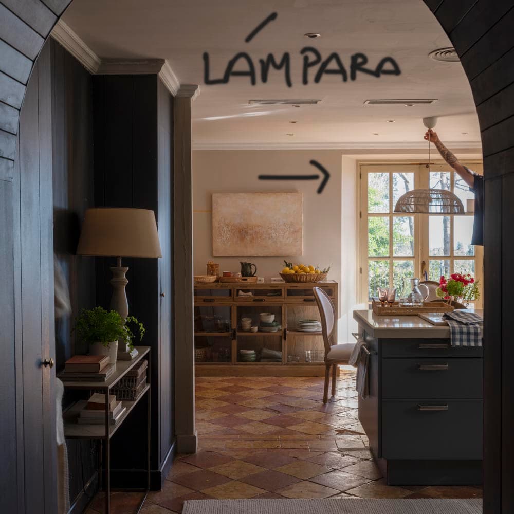 Fotografía de referencia con anotación 'Lámpara' y flecha indicativa, mostrando la vista desde el office a través del arco hacia la cocina con la isla gris-verde, aparador de madera y una lámpara de ratán siendo instalada a la derecha.