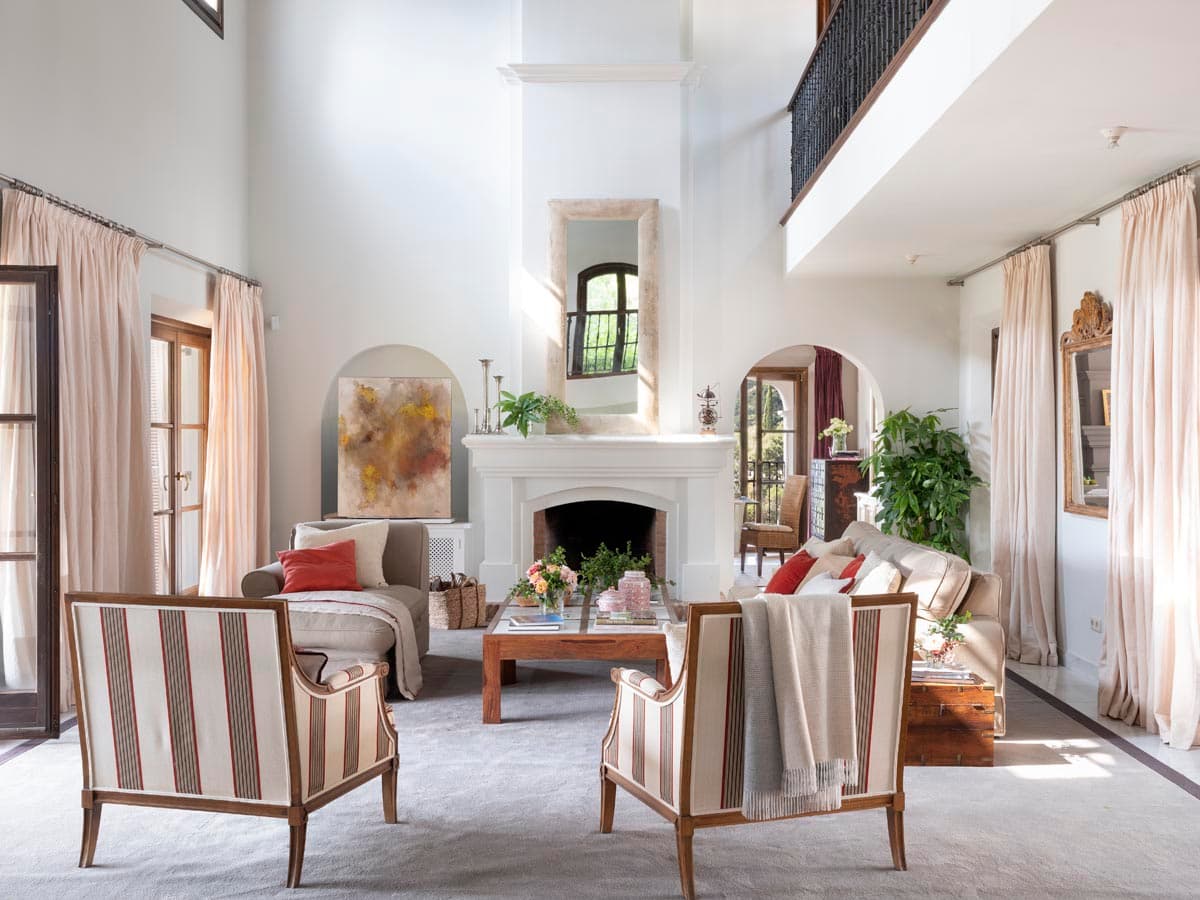 Gran salón de doble altura con cuatro sillones de rayas verticales en tela natural, un sofá gris y una chimenea flanqueada por un cuadro abstracto en ocres y rojos. El mármol del espejo sobre la repisa, los arcos de paso y el balcón superior de hierro forjado subrayan la grandiosidad del espacio.