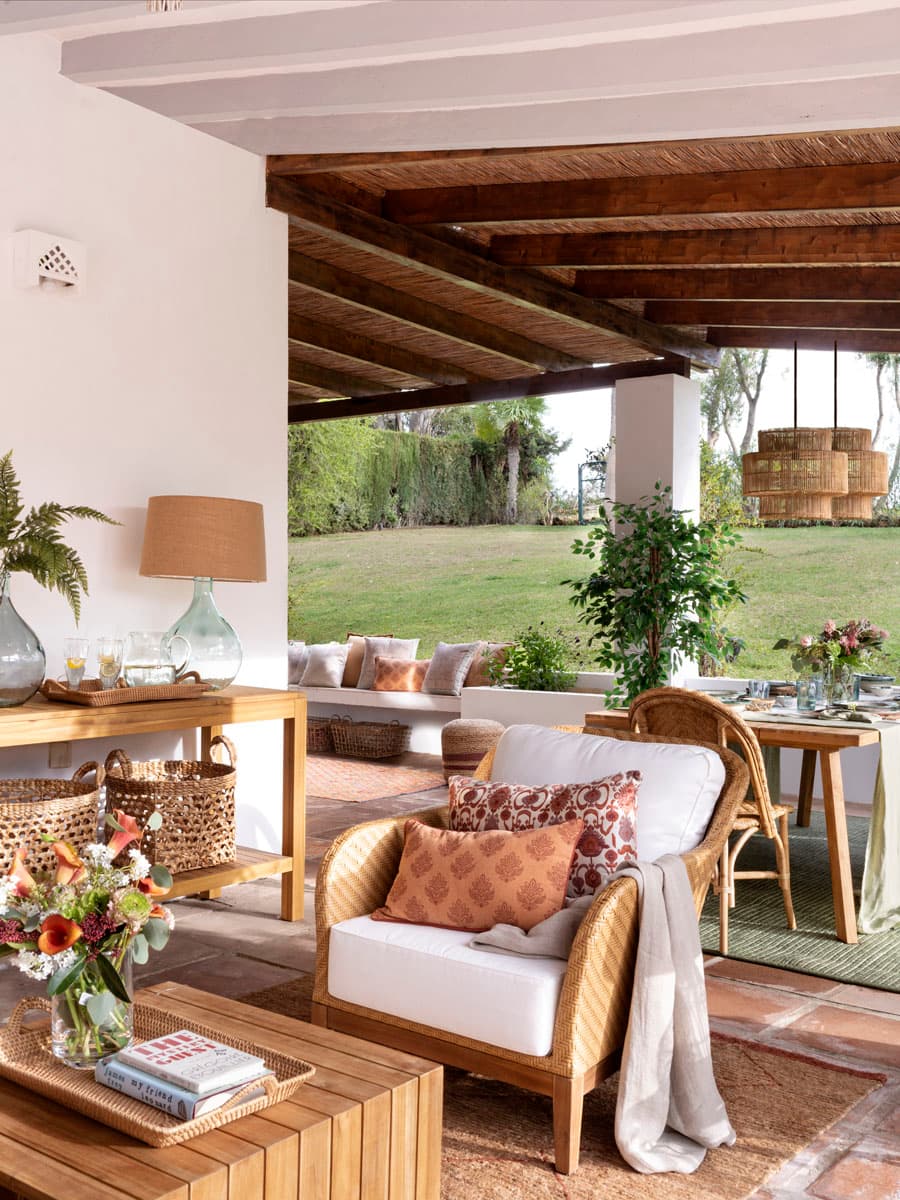 Plano medio del porche con una butaca de ratán tapizada en blanco con cojín de lino estampado en terracota. Detrás, una consola de madera maciza natural con una garrafita de vidrio reciclado verde, copas, una lámpara de sobremesa con pantalla de lino, y un cesto de fibra natural. El fondo se abre hacia la zona de comedor y el jardín con vegetación frondosa. Los elementos reflejan el estilo orgánico, artesanal y mediterráneo del proyecto.
