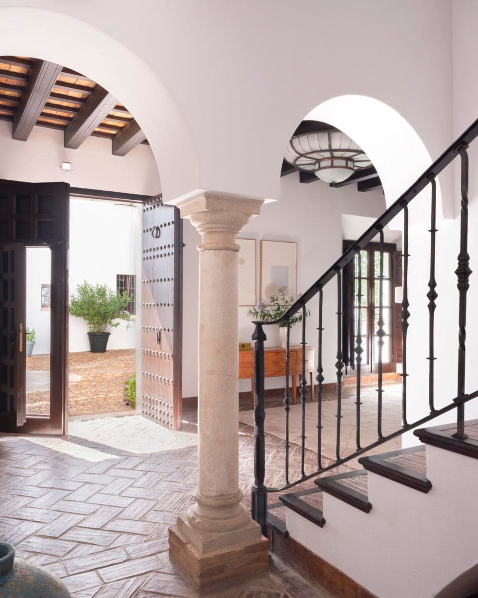 Vestíbulo central con columna de piedra tallada, arcos blancos de herradura, escalera con barandilla de forja negra y puerta principal doble de madera tachonada abierta al patio. Suelo de ladrillo antiguo en espiga.