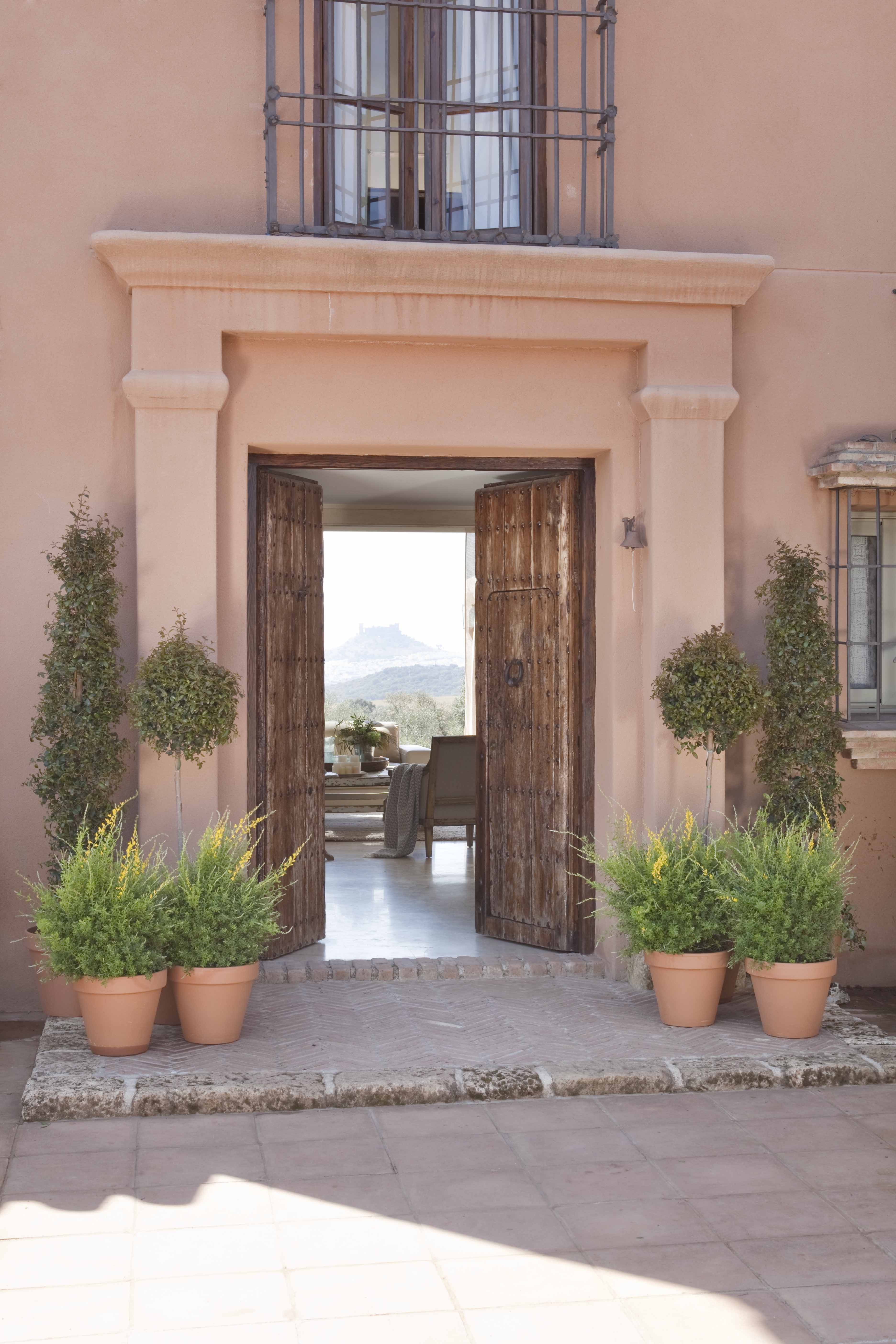 Fachada de entrada en tonos ocre y rosa con portada de piedra tallada y puerta doble de madera antigua con clavos. Jardineras de barro con arbustos podados en forma de columna flanquean la entrada. A través de las puertas abiertas se vislumbra el interior y el paisaje serrano al fondo.