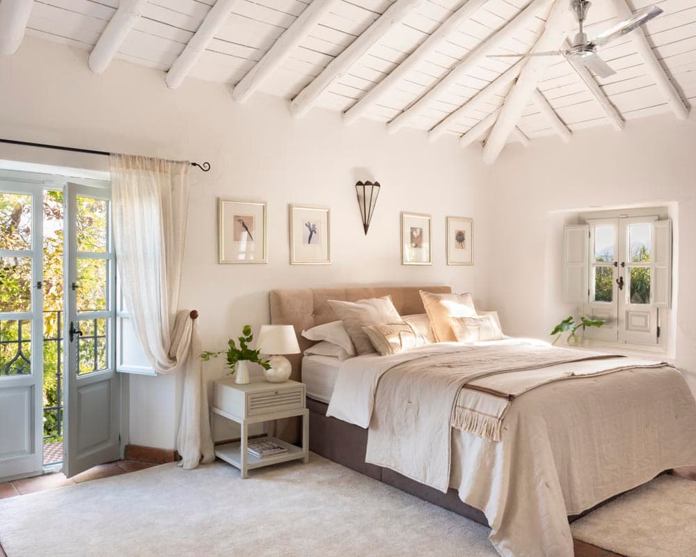 Dormitorio principal amplio con cabecero en lino beige, ropa de cama en tonos naturales y serie de cuadros botánicos sobre la cabecera. Las puertas-ventana con cortinas de lino dan acceso al jardín y un ventilador de techo completa la estancia.