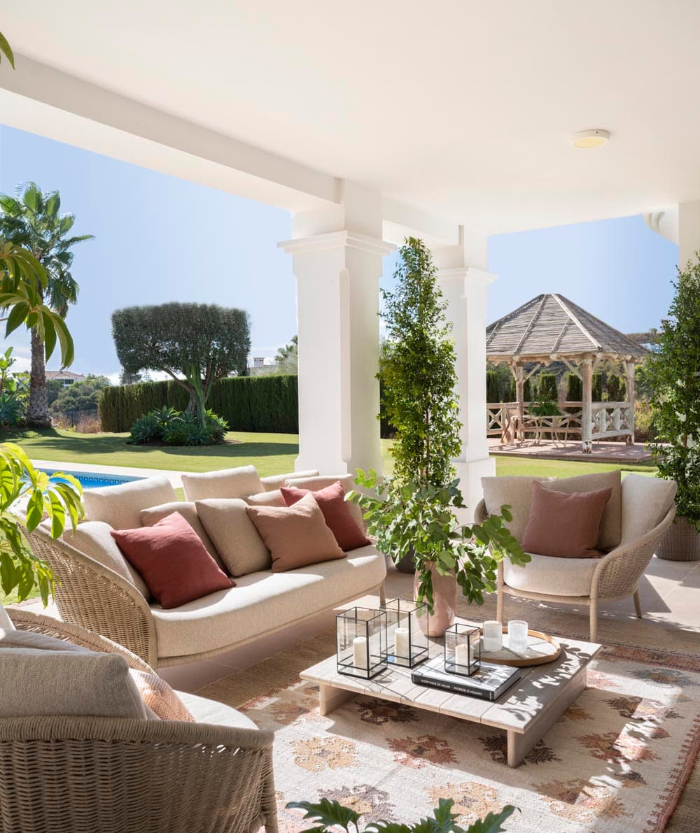 Porche exterior con columnas blancas y amplia zona de estar con sofá de ratán y tejido en beige y cojines en burdeos. Mesa de centro de piedra con farol y vela. El jardín cuidado con piscina azul, palmeras y una pérgola de madera al fondo define el ambiente privilegiado de la Costa del Sol.