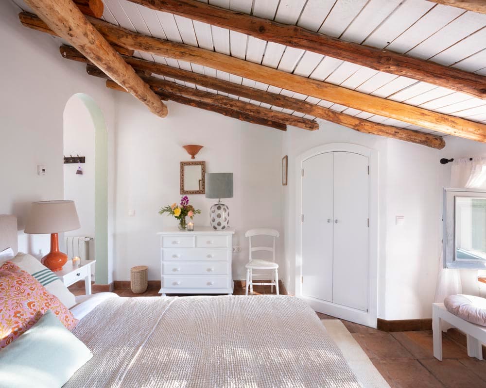 Dormitorio con techumbre de cañas y vigas, chimenea de hierro fundido bajo campana blanca y butaca Luis XV en lino. Una cómoda blanca con espejo dorado, flores de hortensias y cuadros botánicos completan el ambiente romántico.