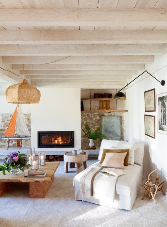 Rincón del salón con chimenea encendida, chaise longue tapizado en blanco con cojín en camel y una lámpara de brazo articulado negro. La estantería de piedra con libros y el cuadro abstracto crean un ambiente acogedor e íntimo junto al fuego.