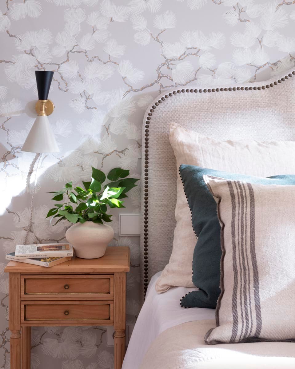 Primer plano de la mesita de noche de madera natural con cajones sobre la que reposan un jarrón de cerámica beis con planta, un libro y un aplique de pared cónico en negro y latón dorado. El cabecero con tachuelas y los cojines en lino y azul petróleo del dormitorio contrastan con el papel pintado de ramas de hojarasca en blanco.