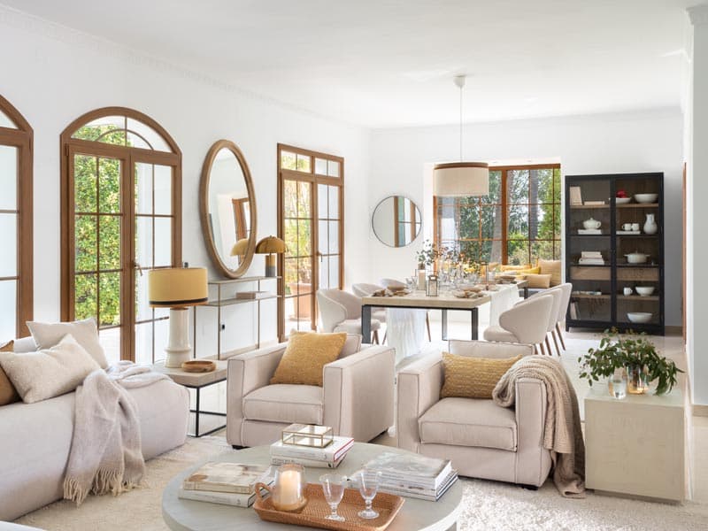 Amplio espacio que integra sala de estar y comedor, con espejos ovales de madera, sillones en beige polvoriento, mesas anidadas y una vitrina oscura que añade profundidad al conjunto.