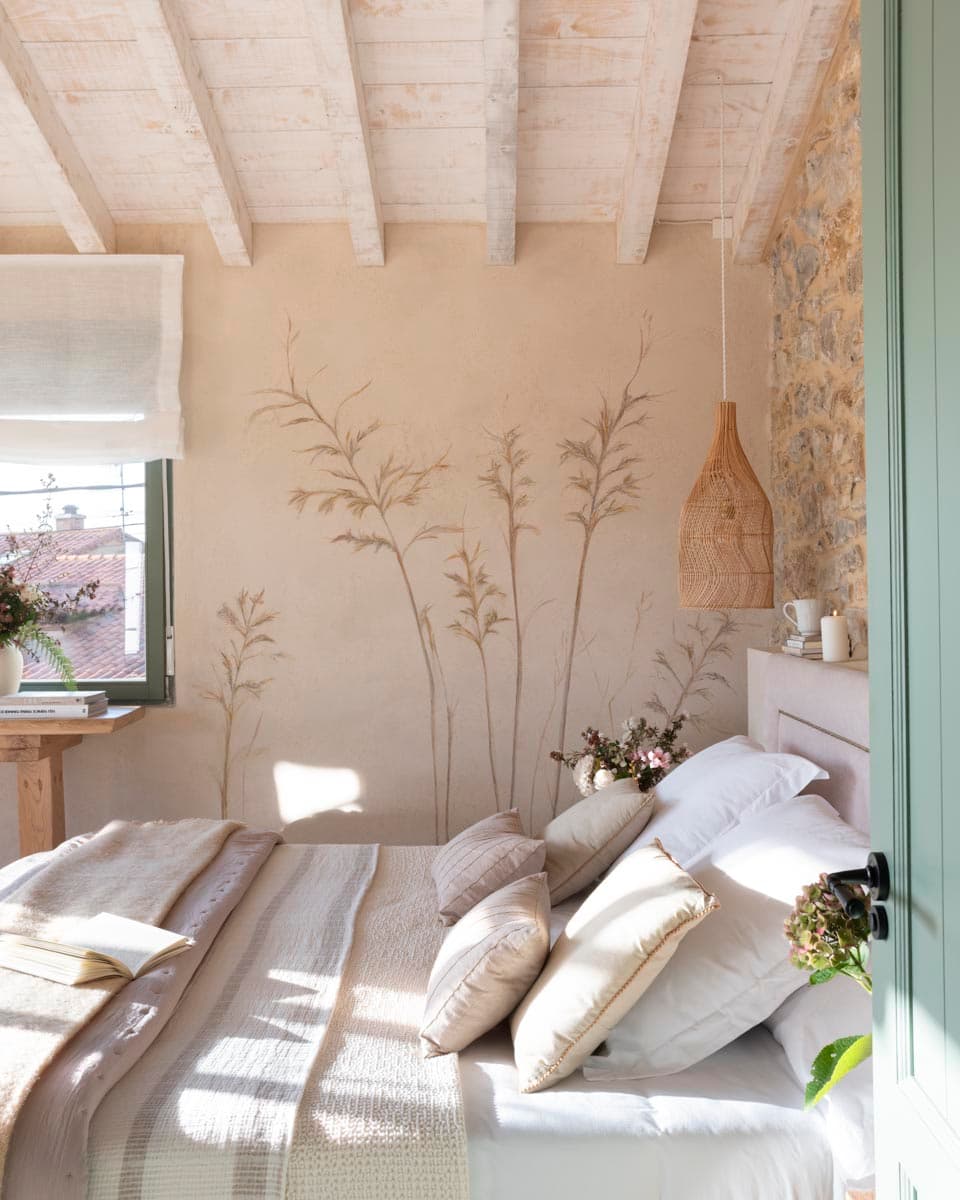Vista frontal del cabecero con un espectacular mural de espigas y gramíneas pintado directamente sobre el enlucido beige, junto a una lámpara colgante de ratán y pared lateral de piedra vista con ventana de marco verde.